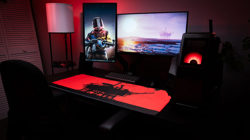 Red and black oil rig deskmat in a dark gaming setup with dual monitors, keyboard, mouse, and red ambient lighting.