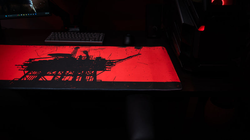 Close-up of a red and black oil rig deskmat with a distressed industrial print in a dark gaming setup with keyboard and mouse.