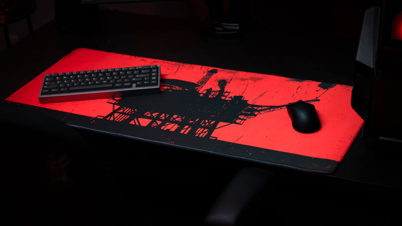 Red and black oil rig gaming deskmat with a distressed industrial design, shown in a dark desk setup with keyboard and mouse.