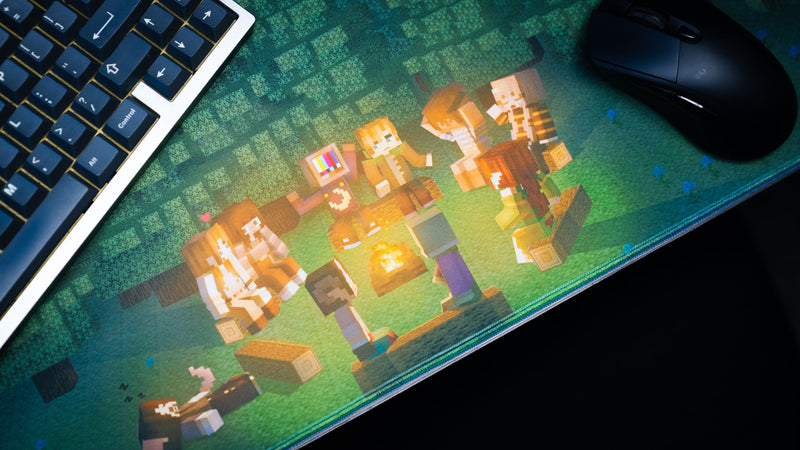 Close-up of the ApolloTV “Adventure Begins” deskmat featuring pixel-style characters gathered around a glowing campfire beside a keyboard and mouse.