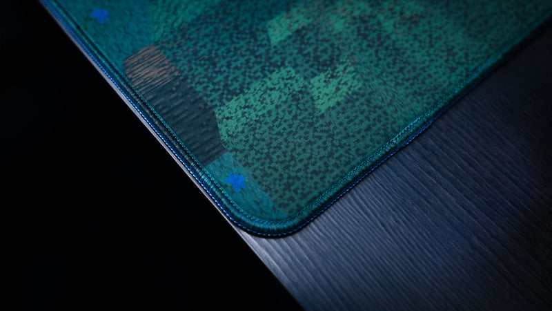 Close-up of the ApolloTV “Adventure Begins” deskmat corner featuring stitched edges, pixel-style forest artwork, and premium fabric texture on a dark desk.
