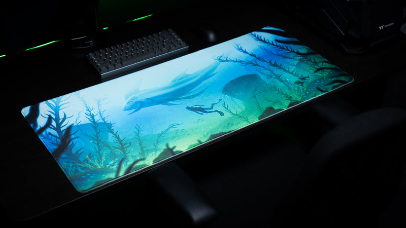 Angled view of Alidove's Lurking Leviathan deskmat showing a sea creature and diver, with a keyboard and PC behind it.