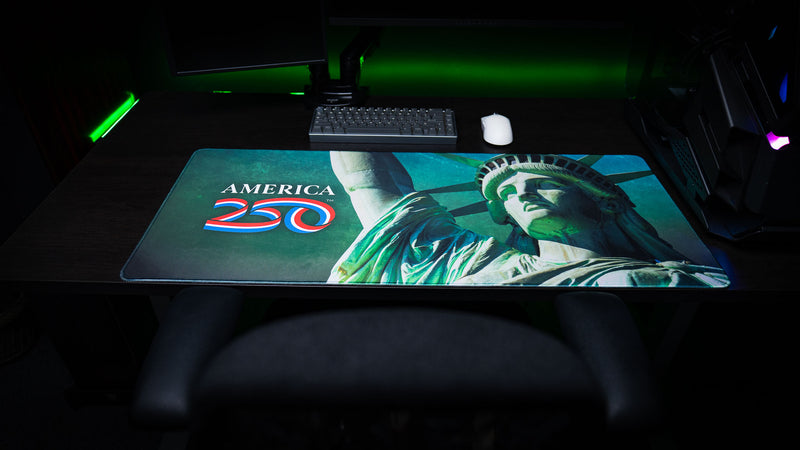 Lifestyle photo of the America 250 Statue of Liberty deskmat displayed on a desktop setup, featuring a bold close-up of the Statue of Liberty, the America 250 logo, premium stitched edges, and a vibrant patriotic design.