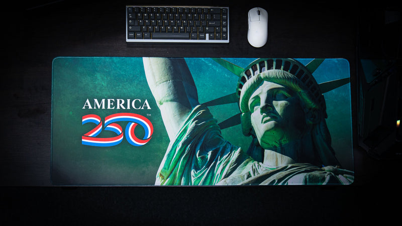 Top-down view of the America 250 Statue of Liberty deskmat featuring a detailed close-up of the Statue of Liberty alongside the America 250 logo on a textured green background with premium stitched edges.