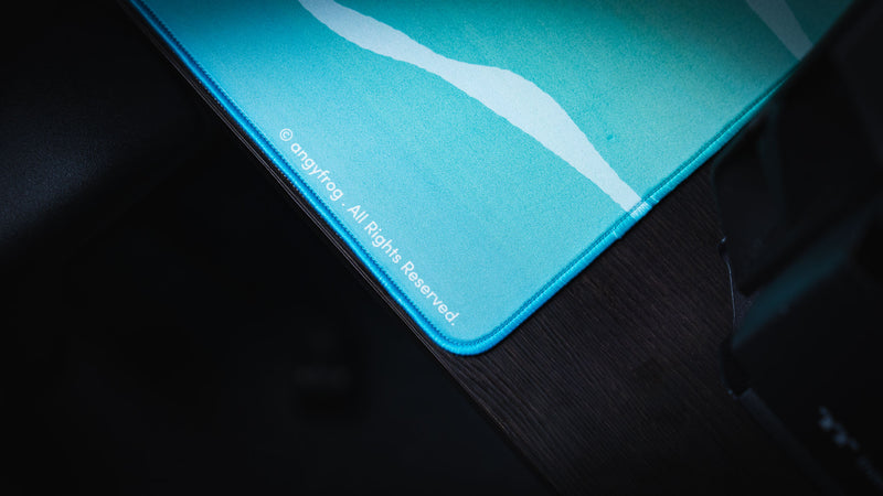 Rounded stitched corner with Angyfrog artist mark on the turquoise Frogs on a Log desk mat