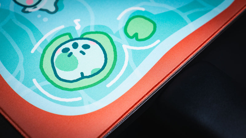 Close-up of a smiling frog face, lily pads, and red stitched edge on Angyfrog The Pond desk mat