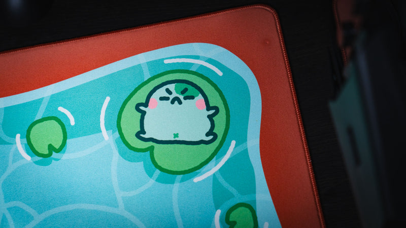 Sleeping frog character on a green lily pad near the corner of the Angyfrog The Pond desk mat