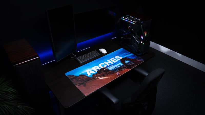 Arches National Park America 250 deskmat displayed in a blue-lit gaming room with Delicate Arch artwork, keyboard, mouse, PC, and dark desk setup.