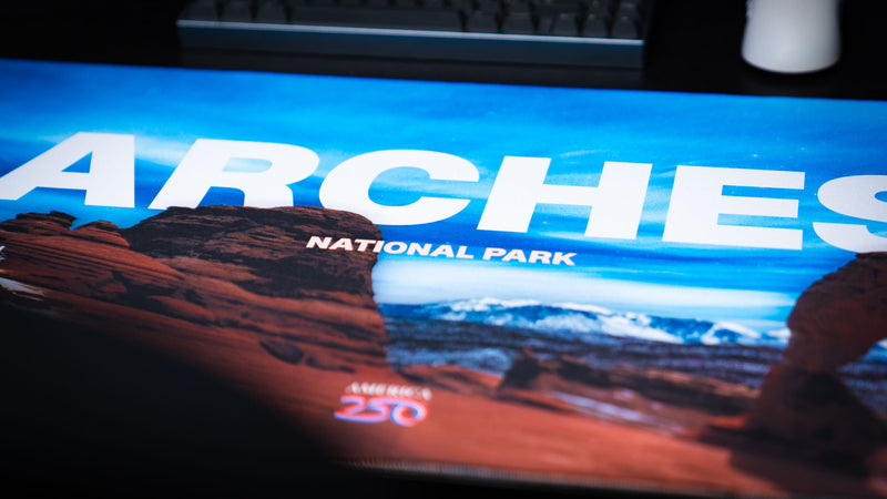 Close-up of the Arches National Park America 250 deskmat showing bold ARches lettering, red rock formations, blue sky artwork, and America 250 logo.