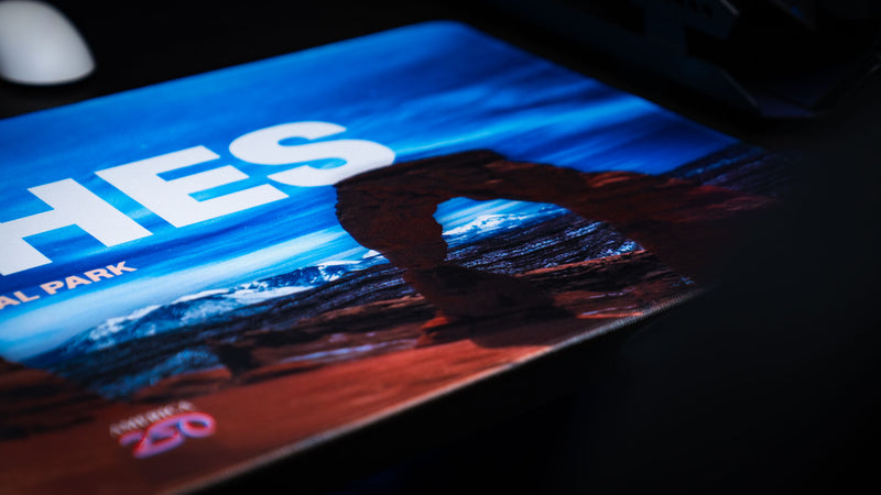 Low-angle close-up of the Arches National Park America 250 deskmat featuring Delicate Arch, bold title text, red rock scenery, and blue sky artwork.