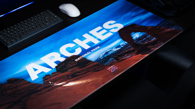 Angled view of the Arches National Park America 250 deskmat featuring Delicate Arch, bold ARches title text, blue sky artwork, keyboard, and mouse.