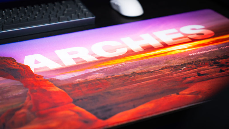 Angled close-up of the Arches National Park premium deskmat featuring Delicate Arch at sunset, vibrant desert colors, and a smooth stitched-edge gaming surface.