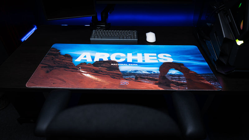 Front view of the Arches National Park America 250 deskmat featuring Delicate Arch, red desert rock formations, bold ARches title text, keyboard, and mouse.