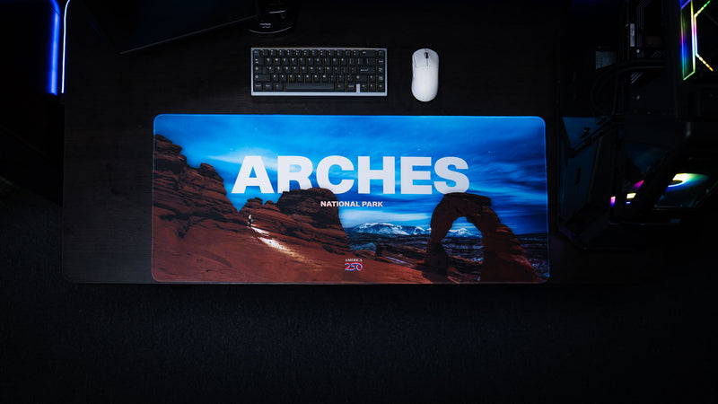 Overhead view of the Arches National Park America 250 deskmat featuring Delicate Arch, bold Arches title text, keyboard, and white mouse.