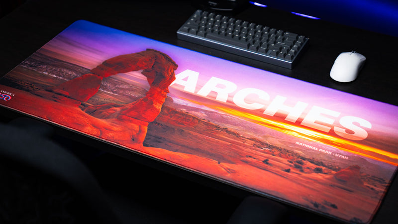 Angled view of the Arches National Park premium deskmat featuring Delicate Arch at sunset, vivid landscape photography, stitched edges, and an extended gaming surface.