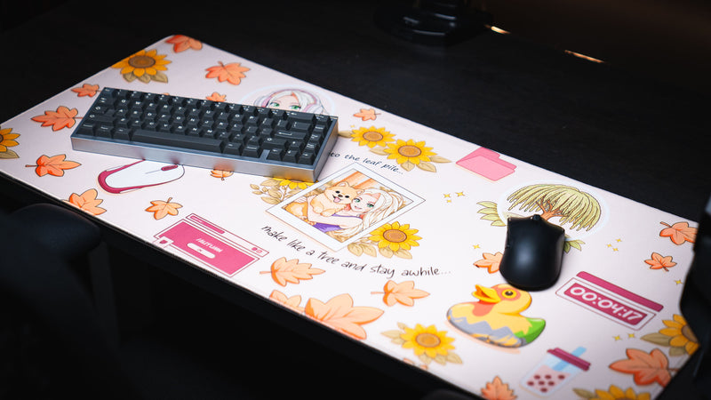 Lifestyle photo of the Cozy Desk deskmat featuring autumn leaves, sunflowers, a cozy Polaroid illustration, colorful rubber duck, pastel desktop icons, and a minimalist gaming keyboard and mouse setup