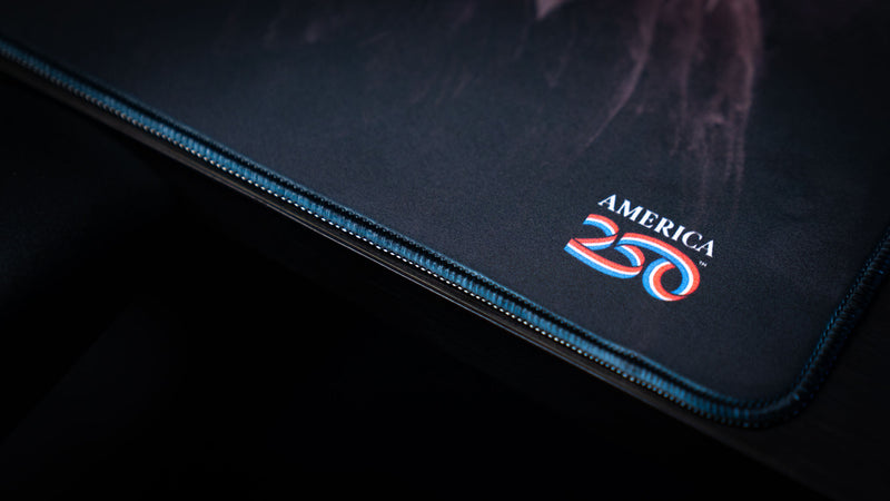 Close-up of the Bald Eagle America 250 deskmat corner showing the America 250 logo, dark patriotic artwork, and premium stitched border.