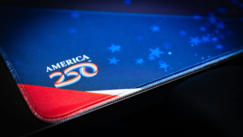 Close-up of the Bald Eagle Blue deskmat corner showing America 250 branding, red white and blue artwork, star details, and stitched edge.