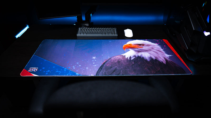 Full-width Bald Eagle Blue America 250 deskmat featuring a bald eagle, blue star background, red accents, keyboard, mouse, and dark desk setup.
