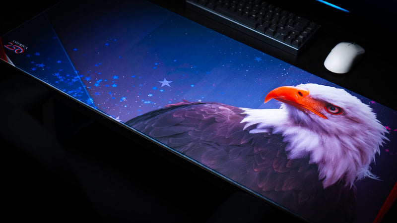 Close-up of the Bald Eagle Blue America 250 deskmat featuring a bald eagle, blue starfield artwork, stitched edge, keyboard, and mouse.