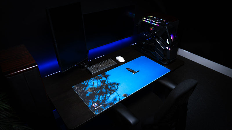Angled lifestyle view of the Bald Eagle in Flight deskmat on a gaming desk, featuring a soaring bald eagle against a vivid blue sky with silhouetted trees from rural Utah as part of the America 250 collection.