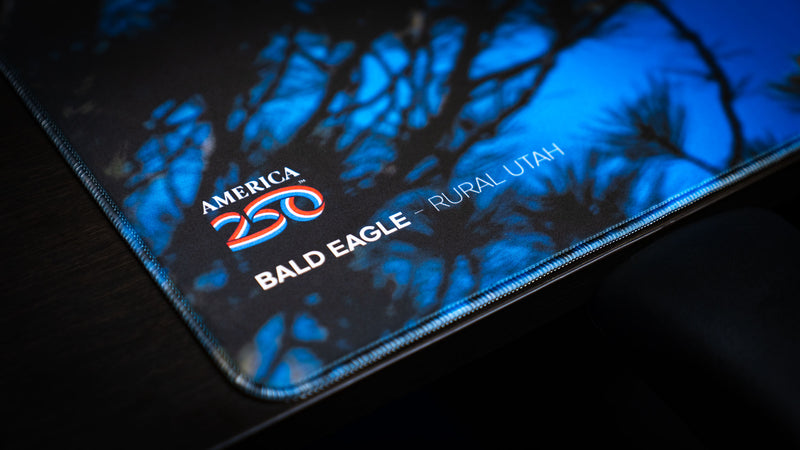 Close-up of the Bald Eagle in Flight deskmat highlighting the America 250 logo, stitched edge, and premium print quality with silhouetted pine branches and a blue Utah sky.
