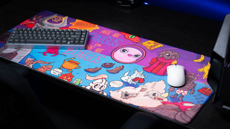 Bingus Bongus deskmat shown with a mechanical keyboard and white gaming mouse, highlighting the colorful artwork and full-size desk coverage.