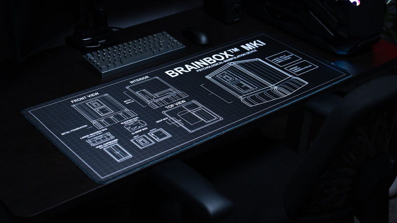 Angled view of the BRAINBOX MK1 deskmat featuring detailed Rust-inspired blueprint schematics, base layouts, and technical building diagrams on a premium gaming deskmat.