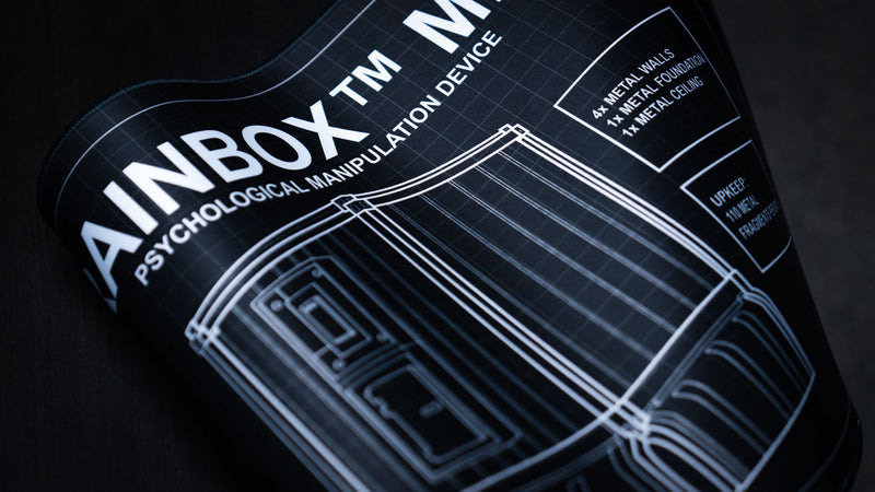 Close-up of the BRAINBOX MK1 deskmat featuring a detailed Rust metal base blueprint with technical schematic artwork and construction specifications.