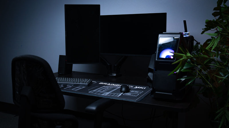 BRAINBOX MK1 deskmat featured in a complete gaming battlestation with dual monitors, mechanical keyboard, and Rust-inspired blueprint artwork.
