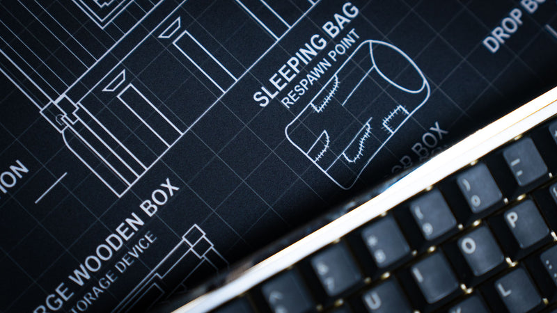 Close-up of the BRAINBOX MK1 deskmat featuring a Rust-inspired sleeping bag respawn point blueprint and detailed schematic artwork.