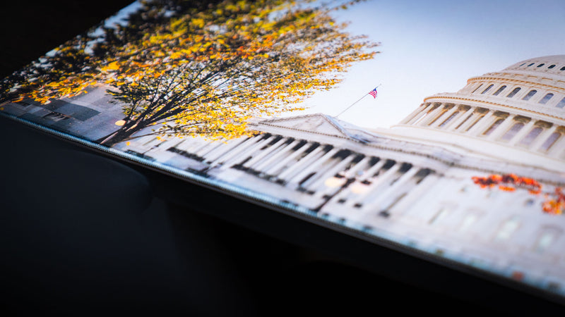 Angled close-up of the Capitol Building deskmat featuring the United States Capitol, American flag, colorful fall trees, and premium stitched edges on a large gaming desk mat.