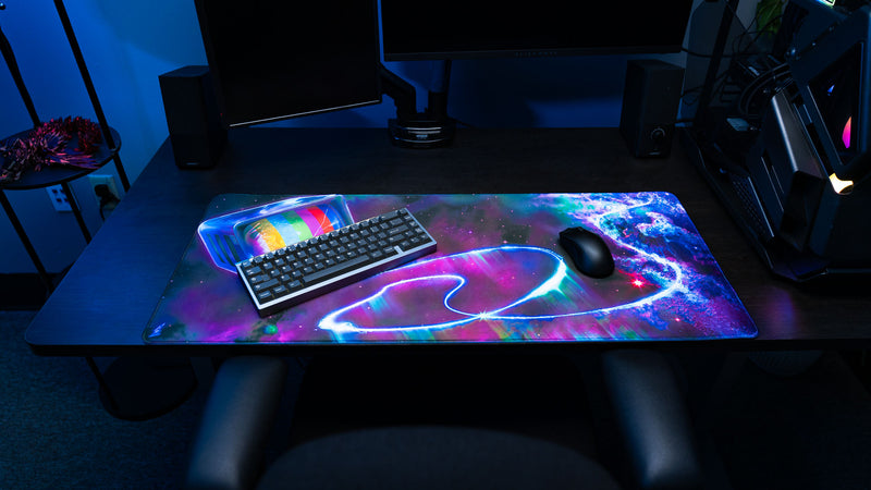 ApolloTV Cosmic TV XXL deskmat displayed in a gaming setup with a retro rainbow television design, glowing cosmic trails, and vibrant nebula artwork.