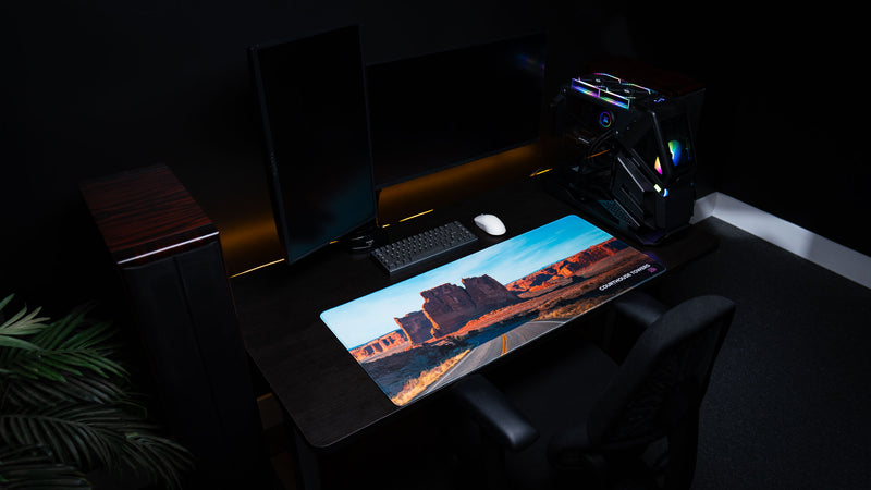 Courthouse Towers America 250 deskmat displayed in a dark ambient gaming setup with Arches National Park rock towers, desert road artwork, keyboard, mouse, and RGB PC.
