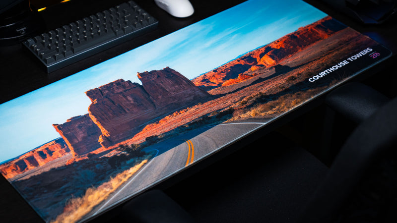 Angled view of the Courthouse Towers America 250 deskmat featuring Arches National Park red rock towers, desert road artwork, keyboard, and white mouse.