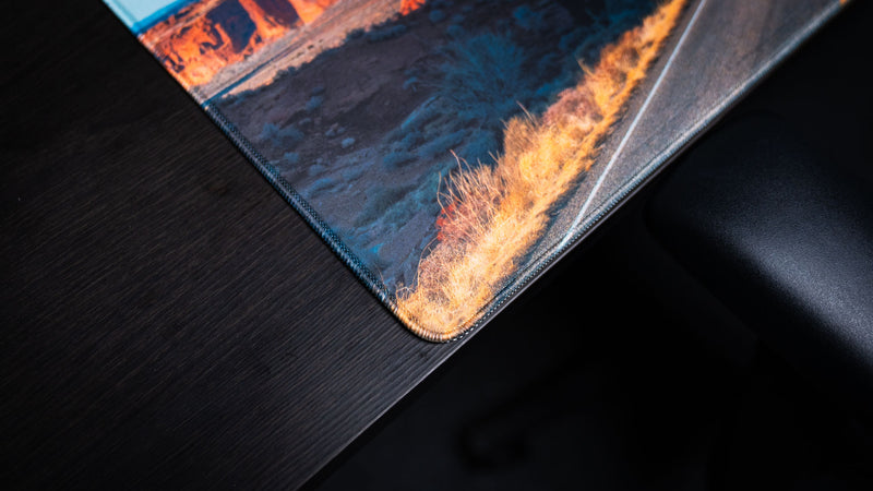 Close-up of the Courthouse Towers America 250 deskmat corner showing desert grass artwork, road detail, fabric texture, and stitched edge.