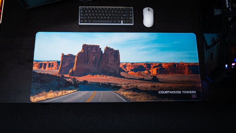 Overhead view of the Courthouse Towers America 250 deskmat featuring Arches National Park rock towers, desert road scenery, keyboard, and white mouse.