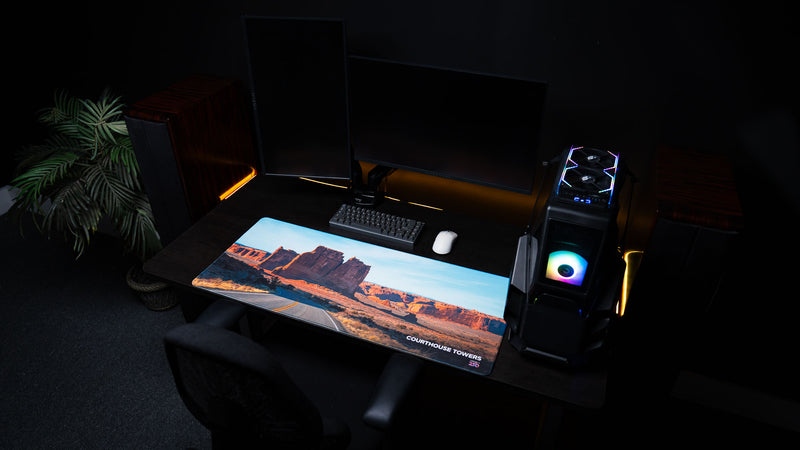 Courthouse Towers America 250 deskmat displayed in an RGB PC setup with Arches National Park sandstone towers, desert road scenery, keyboard, and mouse.