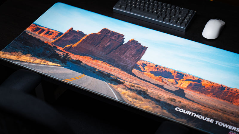 Courthouse Towers America 250 deskmat shown with keyboard and mouse, featuring Arches National Park sandstone towers, desert highway artwork, and blue sky.