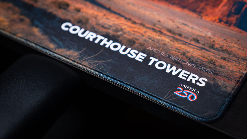 Close-up of the Courthouse Towers America 250 deskmat showing the printed title, Arches National Park text, desert artwork, and stitched edge.