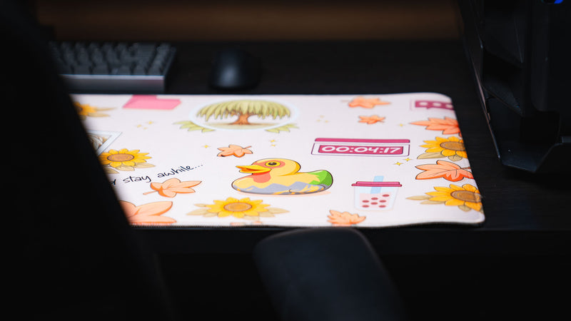 Close-up of the Cozy Desk deskmat featuring a colorful rubber duck, autumn leaves, sunflowers, a willow tree illustration, and cozy pastel desktop-themed artwork.