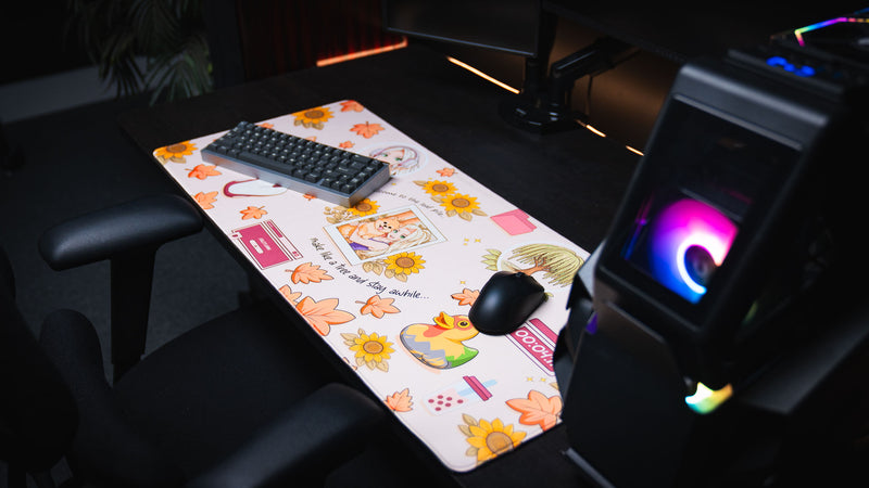 Angled view of the Cozy Desk deskmat in a gaming setup featuring autumn leaves, sunflowers, a Polaroid illustration, colorful rubber duck, pastel desktop icons, and a cozy seasonal aesthetic.