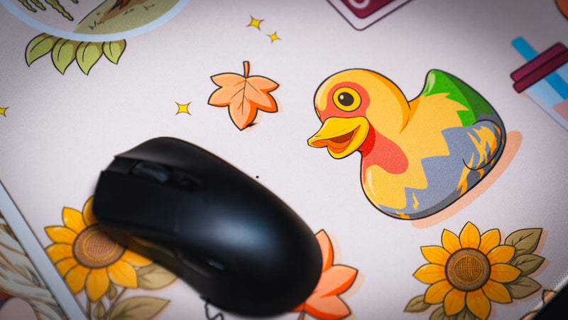 Close-up of the Cozy Desk deskmat featuring a colorful rubber duck illustration surrounded by autumn leaves, sunflowers, and a gaming mouse