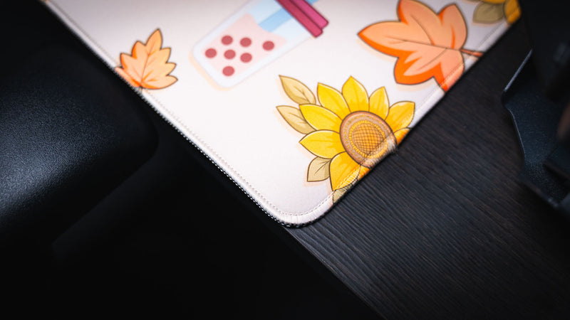 Close-up of the Cozy Desk deskmat showcasing premium stitched edges with sunflower artwork, autumn leaves, and a pastel bubble tea illustration in the corner.