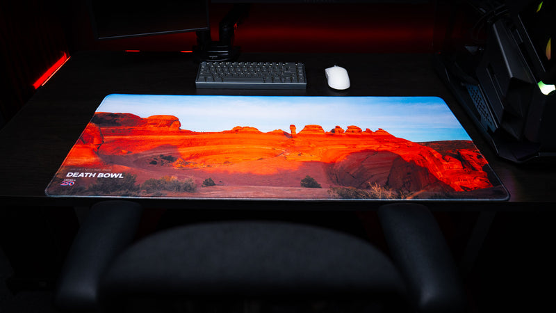 Death Bowl deskmat displayed in a premium gaming setup, showcasing the sweeping red sandstone basin of Arches National Park with vibrant desert colors and stitched edges.