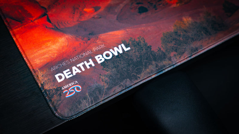 Close-up of the Death Bowl deskmat featuring the Arches National Park title, America 250 logo, premium stitched edges, and vibrant red rock photography.