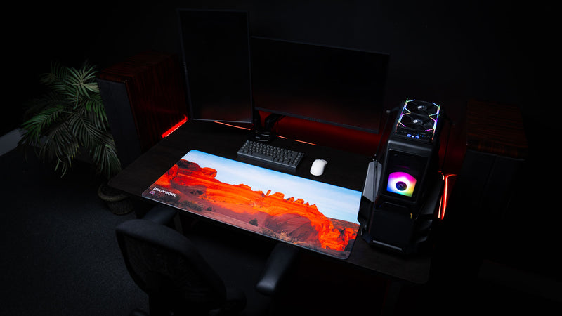 Death Bowl deskmat displayed in a modern gaming workstation, featuring the iconic red sandstone basin of Arches National Park with vivid sunset colors.