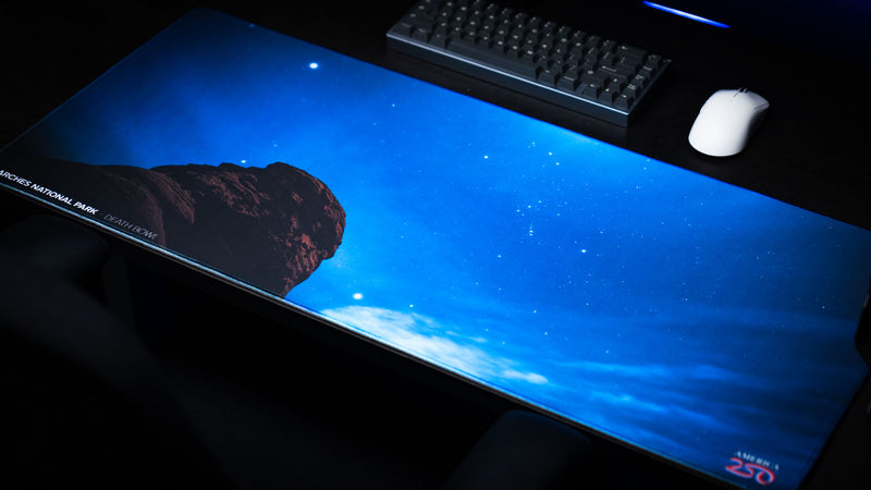 Angled view of the Death Bowl Nightsky deskmat featuring a brilliant star-filled sky over the iconic Arches National Park rock formation on a premium gaming desk setup