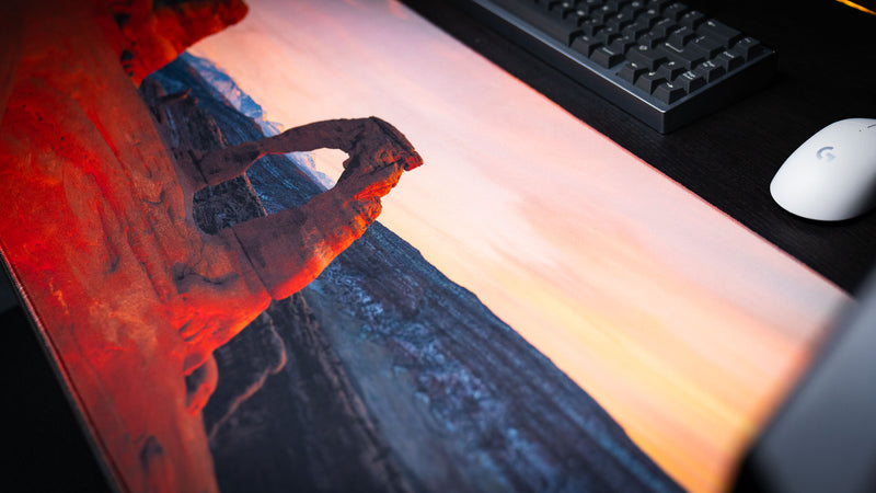 Close-up of the Delicate Arch premium deskmat in a gaming setup, showcasing iconic Arches National Park photography, vibrant sunrise colors, and a smooth stitched-edge surface.