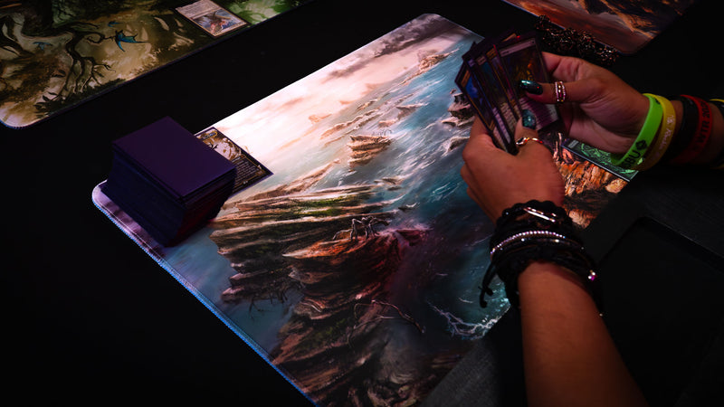 Desolate Island playmat in use during a trading card game, featuring rocky coastal cliffs, scattered islands, and crashing ocean waves.